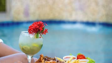 A wicker table with a plate of food and a drink by a pool.