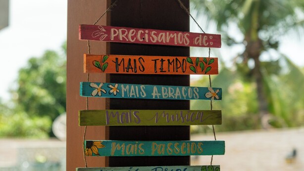A wooden sign with multiple colorful wooden signs hanging from a pole, each with a different phrase in Portuguese. - Bem Bahia Hotel (Porto Seguro)