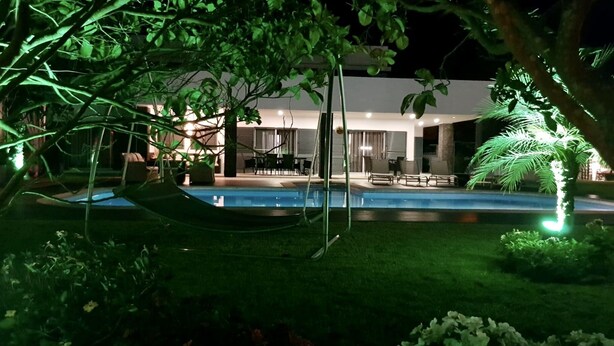 En nattbild av ett modernt hus med en swimmingpool, omgivet av frodig grönska och en hängmatta.