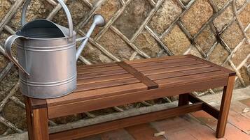 A wooden bench with a metal watering can placed on it, set against a stone wall with a diamond pattern.
