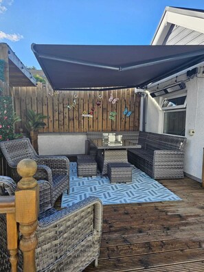 Ein überdachter Patio-Bereich mit Korbmöbeln, einem gemusterten Teppich und dekorativen Wandgestaltungen.
