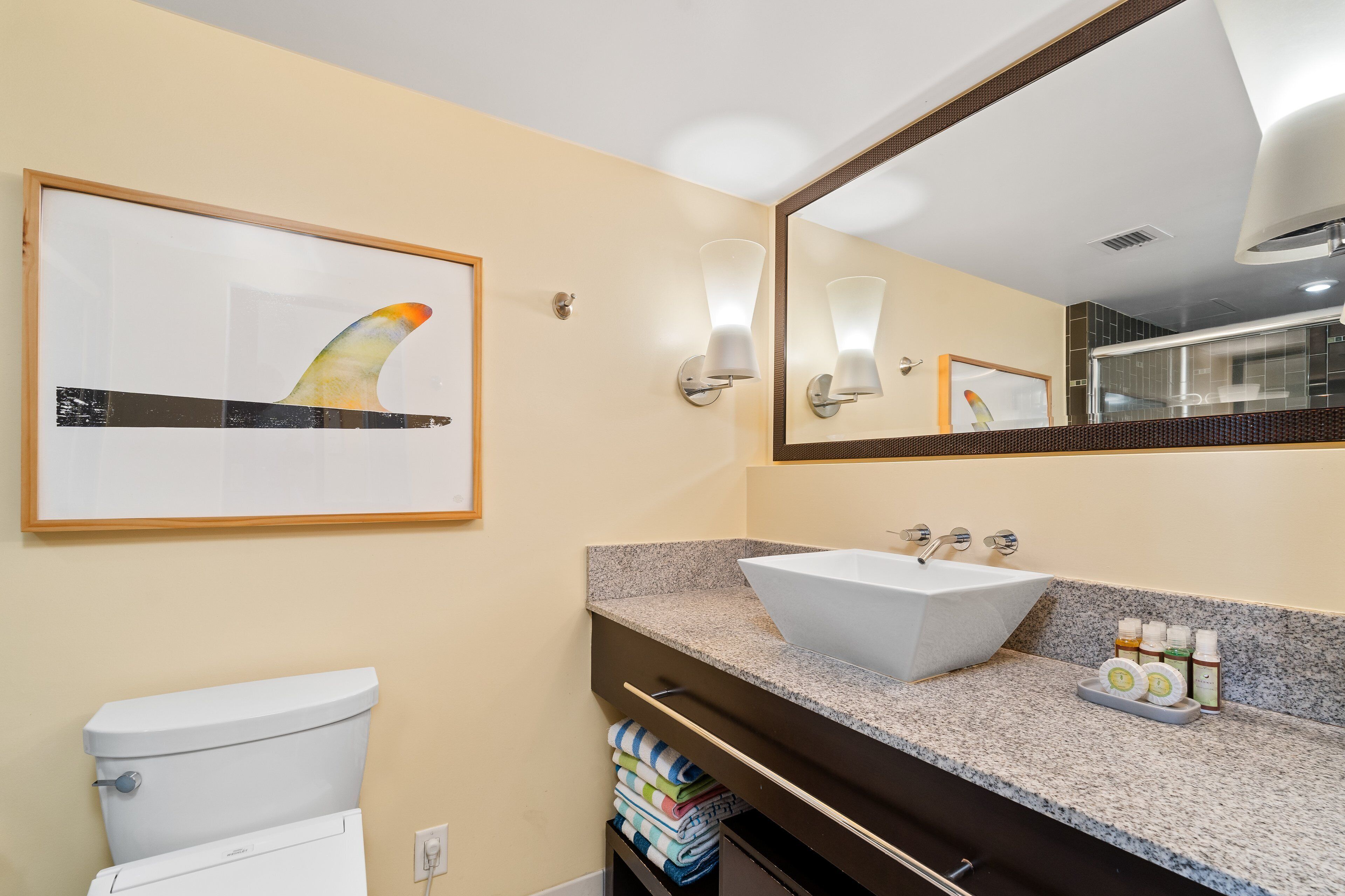 Une salle de bain avec un lavabo blanc, une œuvre d’art représentant une nageoire de requin encadrée, un grand miroir et un porte-serviettes avec des serviettes pliées.