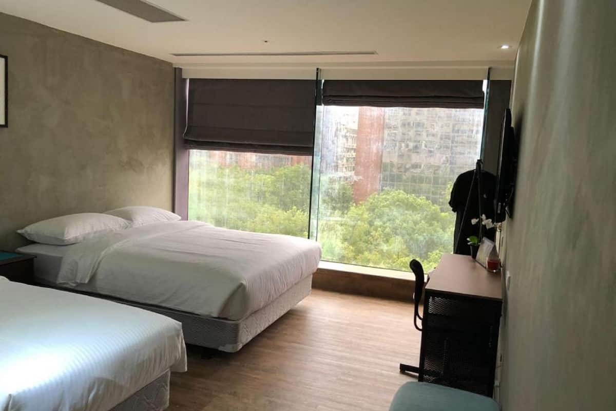 Een slaapkamer met twee bedden, een bureau en een groot raam met uitzicht op bomen en gebouwen.