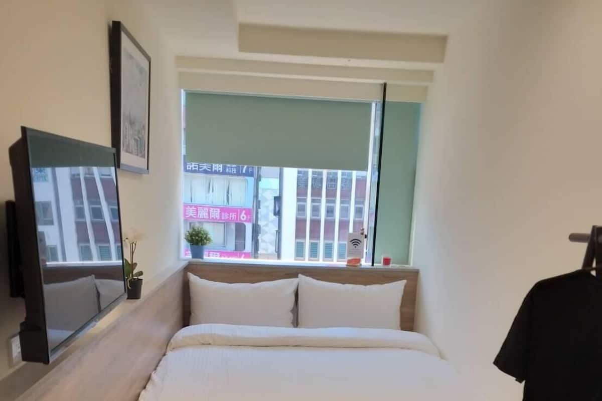Een slaapkamer met een bed, een klein krukje, een televisie, een raam en uitzicht op de straat.