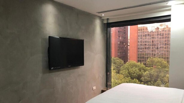Un dormitori amb televisió de pantalla plana penjada a la paret, un llit gran amb roba de llit blanca, i vista de gratacels i arbres per la finestra.
