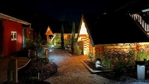 Un sendero de piedra conduce a una cabaña de madera con techo inclinado, rodeada de jardines ajardinados y un edificio rojo.