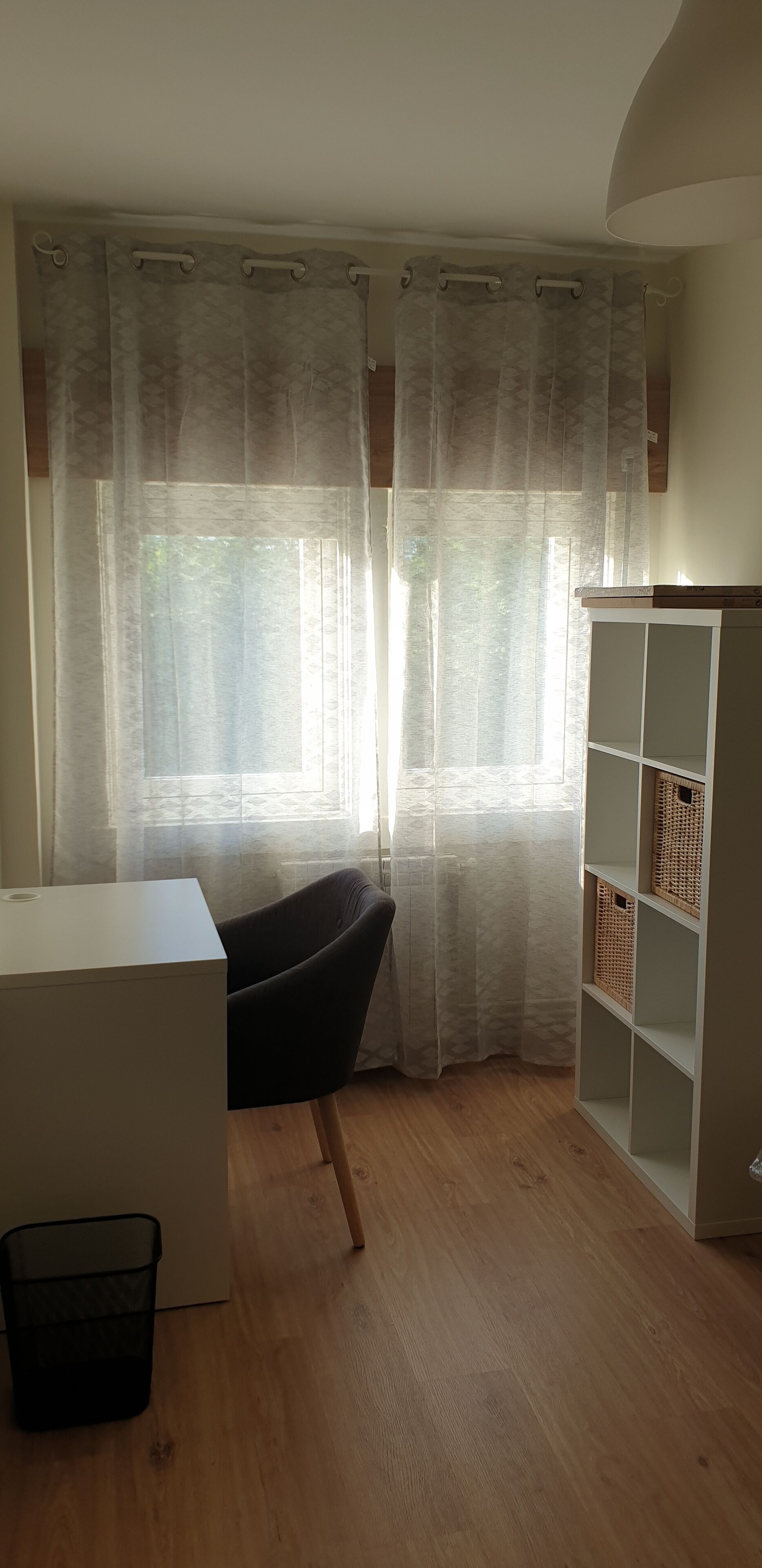 Una habitación con piso de madera, un escritorio blanco, una silla negra, un estante blanco y cortinas translúcidas.