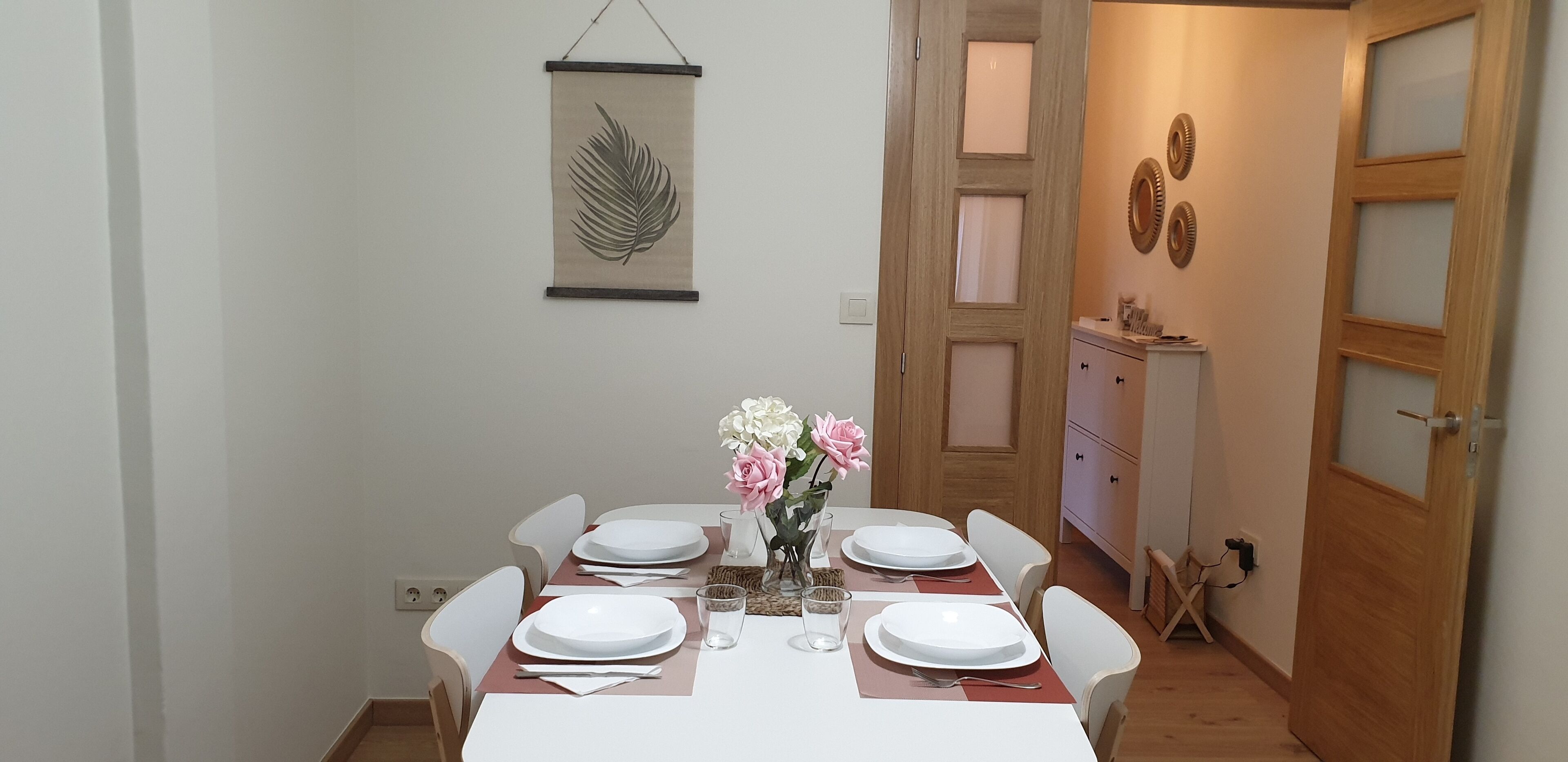 Un comedor con una mesa dispuesta para cuatro, sillas blancas, un jarrón con flores y una obra de arte colgada en la pared.