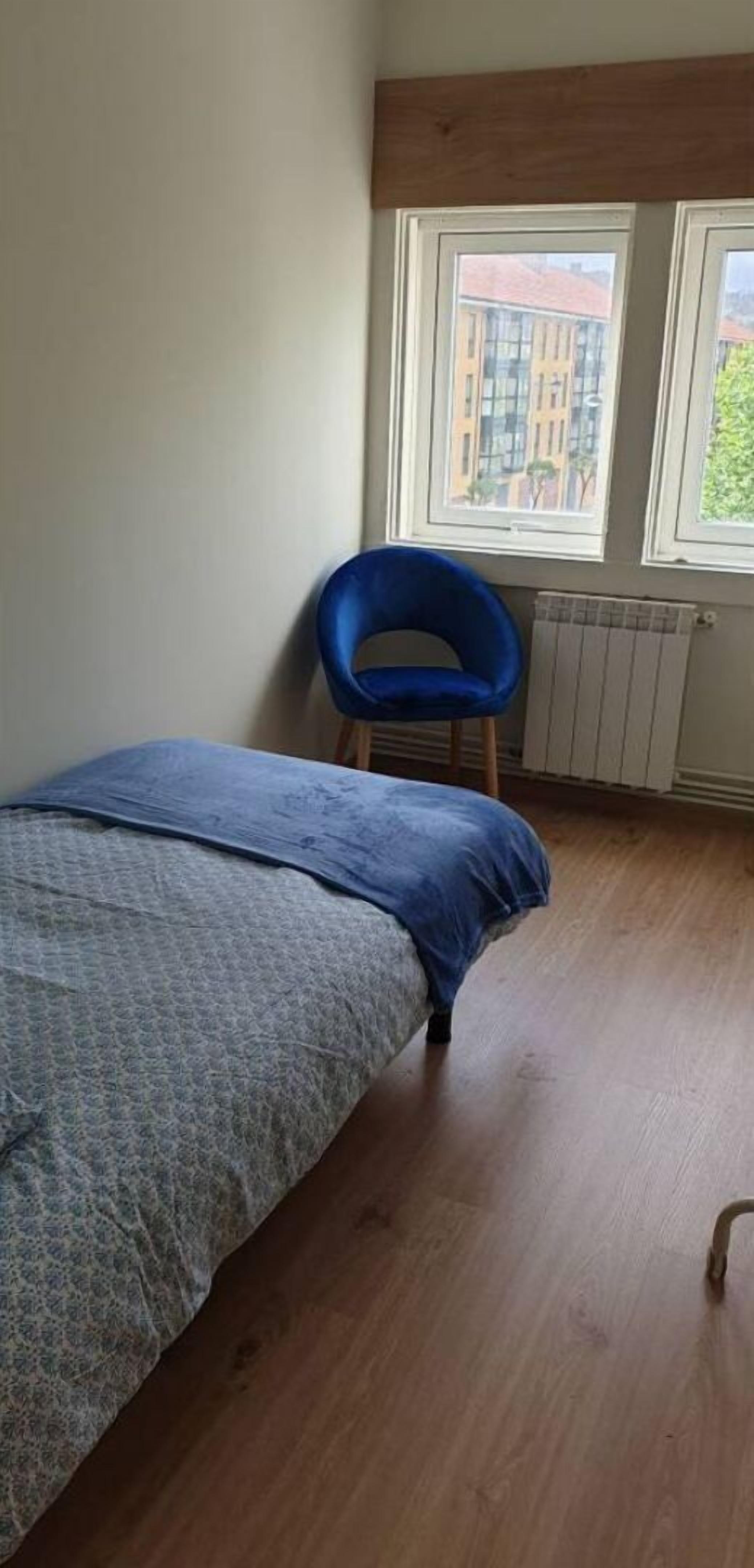 Un dormitorio con una cama, una silla azul y un ventanal con vistas a edificios.