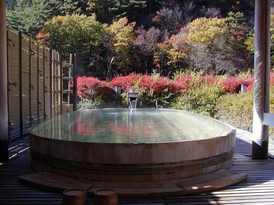 A hot spring bath surrounded by a wooden deck, with a backdrop of colorful foliage and a bamboo fence.