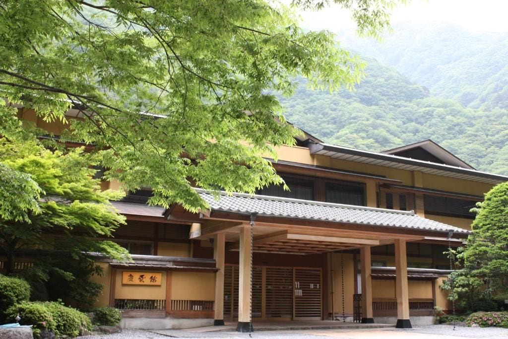 Edificio japonés tradicional con un porche cubierto, rodeado de exuberante vegetación y colinas al fondo.