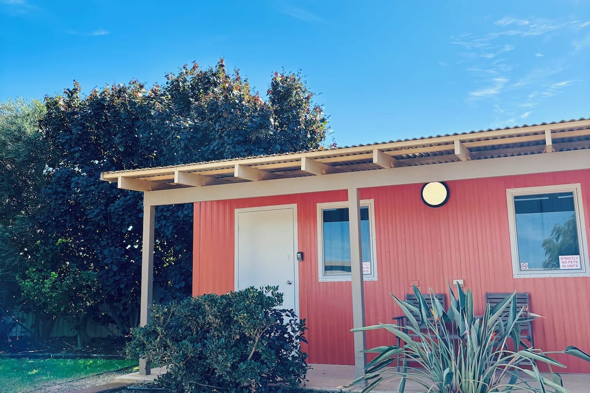 Un edificio rojo con puerta y ventanas blancas, rodeado de follaje y un cielo azul despejado.