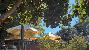 Eine Terrasse mit Sitzgelegenheiten im Freien unter gestreiften Sonnenschirmen, umgeben von Grünpflanzen und blühenden Blumen.