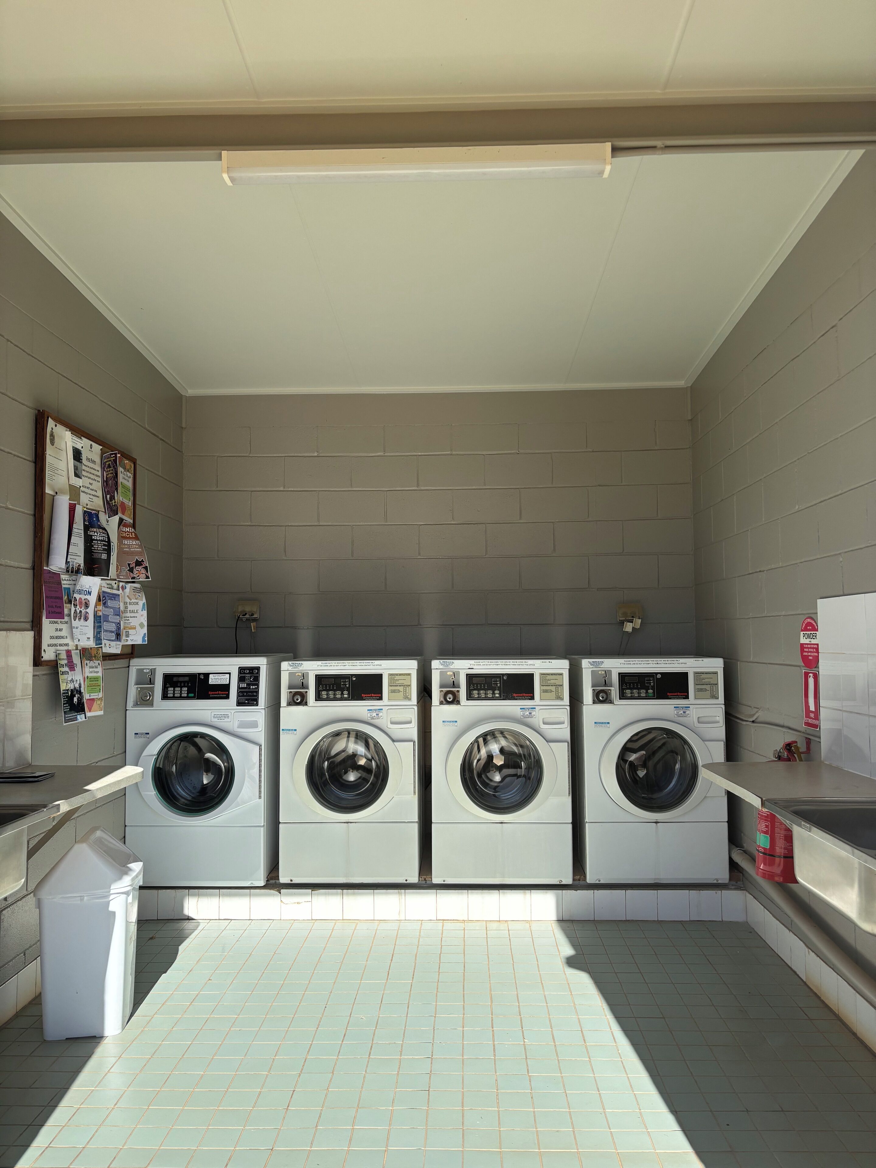 A laundry room with three stacked front-loading washing machines, a bulletin board with various notices, and a tiled floor.