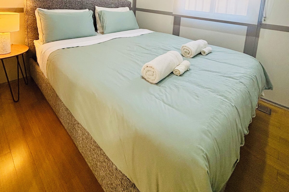 Una cama bien hecha con cubrecama y almohadas verde claro, una mesita de noche con lámpara y una ventana con persianas.