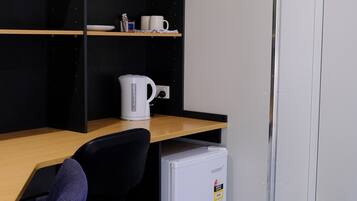 A small office space with a desk, a chair, a white kettle, and a compact refrigerator.