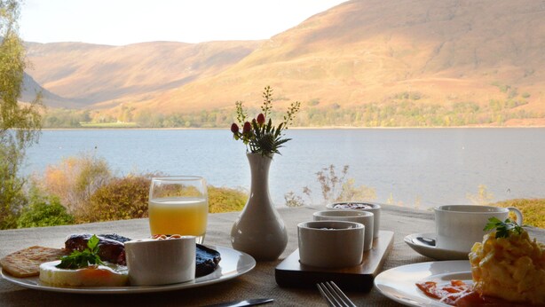 A table set with breakfast items, a vase with flowers, and a view of a lake and hills. - Torlinnhe Guesthouse (Fort William)