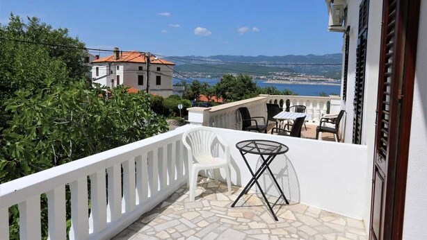 A balcony with a white railing, a small table, and a chair, overlooking a scenic view of a body of water and distant buildings. - Apartment Ljustina (Silo - island Krk)