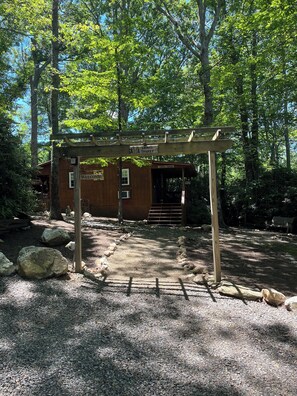 Une cabane en bois avec une pancarte « Bienvenue », entourée d’arbres et de chemins de gravier.