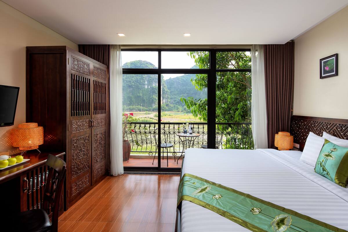 A hotel room with a large bed, a TV, a dining table with chairs, and a balcony with a view of greenery and hills.