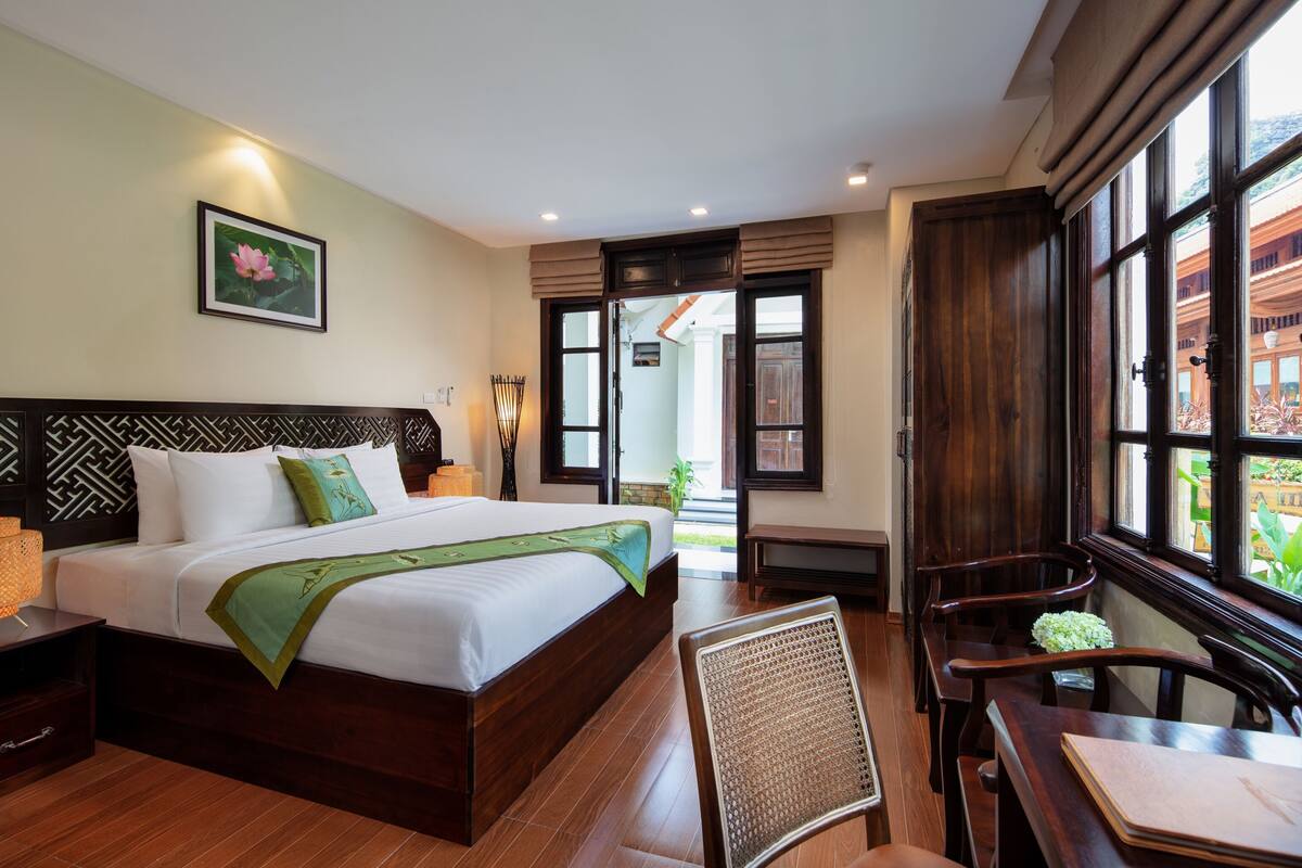 A hotel room with a large bed, a desk, and a chair. There is a window with a view of the outside.