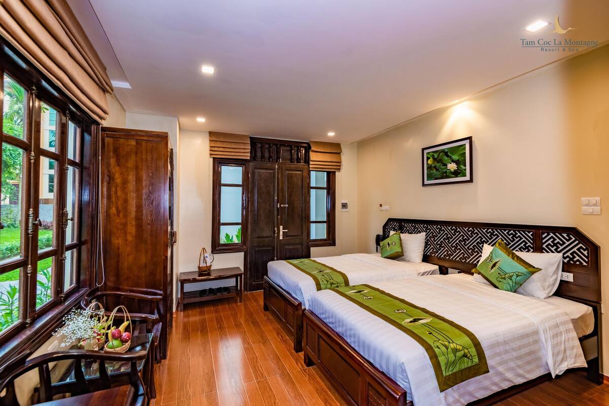 A hotel room with two beds, wooden furniture, a dining table with fruit, and large windows.