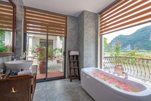 A modern bathroom with a large bathtub, a sink, and a wooden vanity. There is a view of a balcony with flowers and a mountainous landscape.