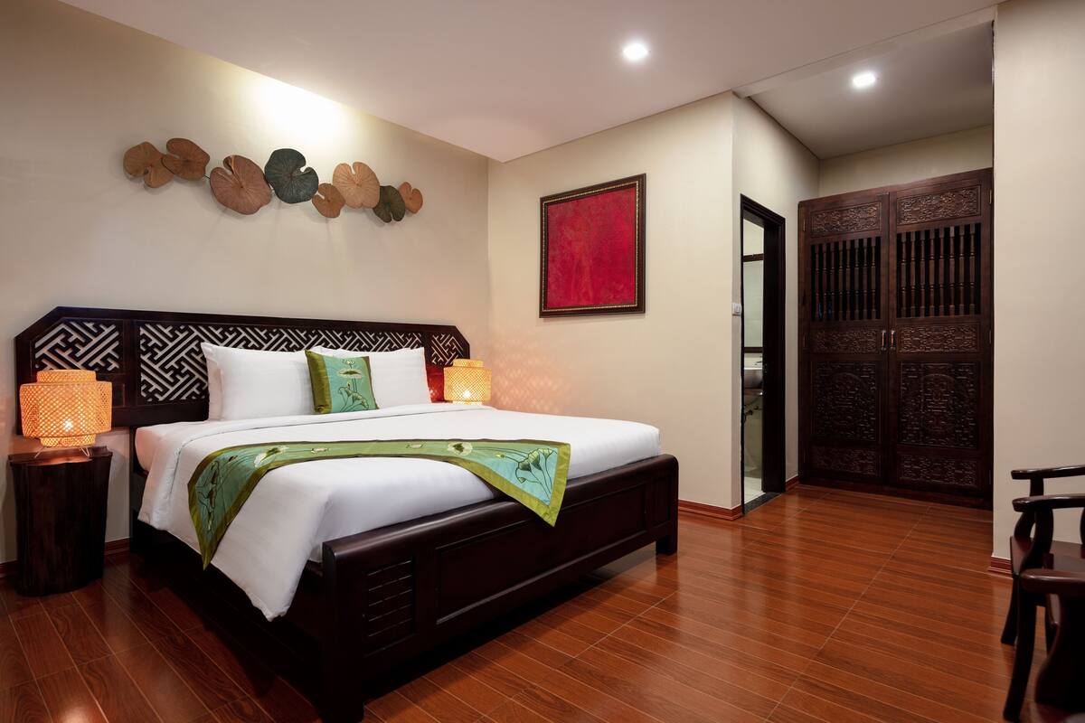 A spacious bedroom with a large bed, wooden floor, decorative wall art, and a traditional wooden wardrobe.