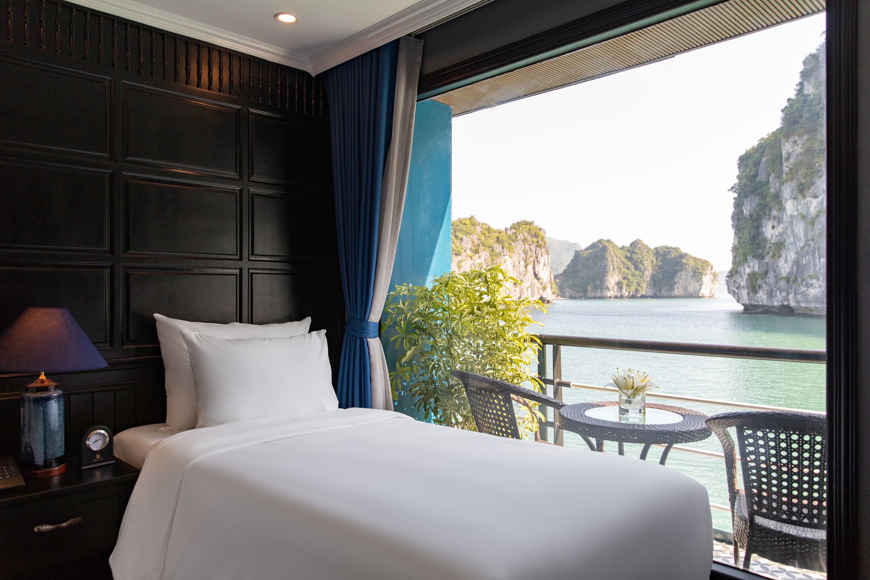 A hotel room with a bed, a nightstand, a table with a chair, and a view of the sea and cliffs.