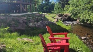 Une chaise Adirondack rouge, placée près d’un ruisseau, avec une terrasse en bois et un bâtiment en arrière-plan.