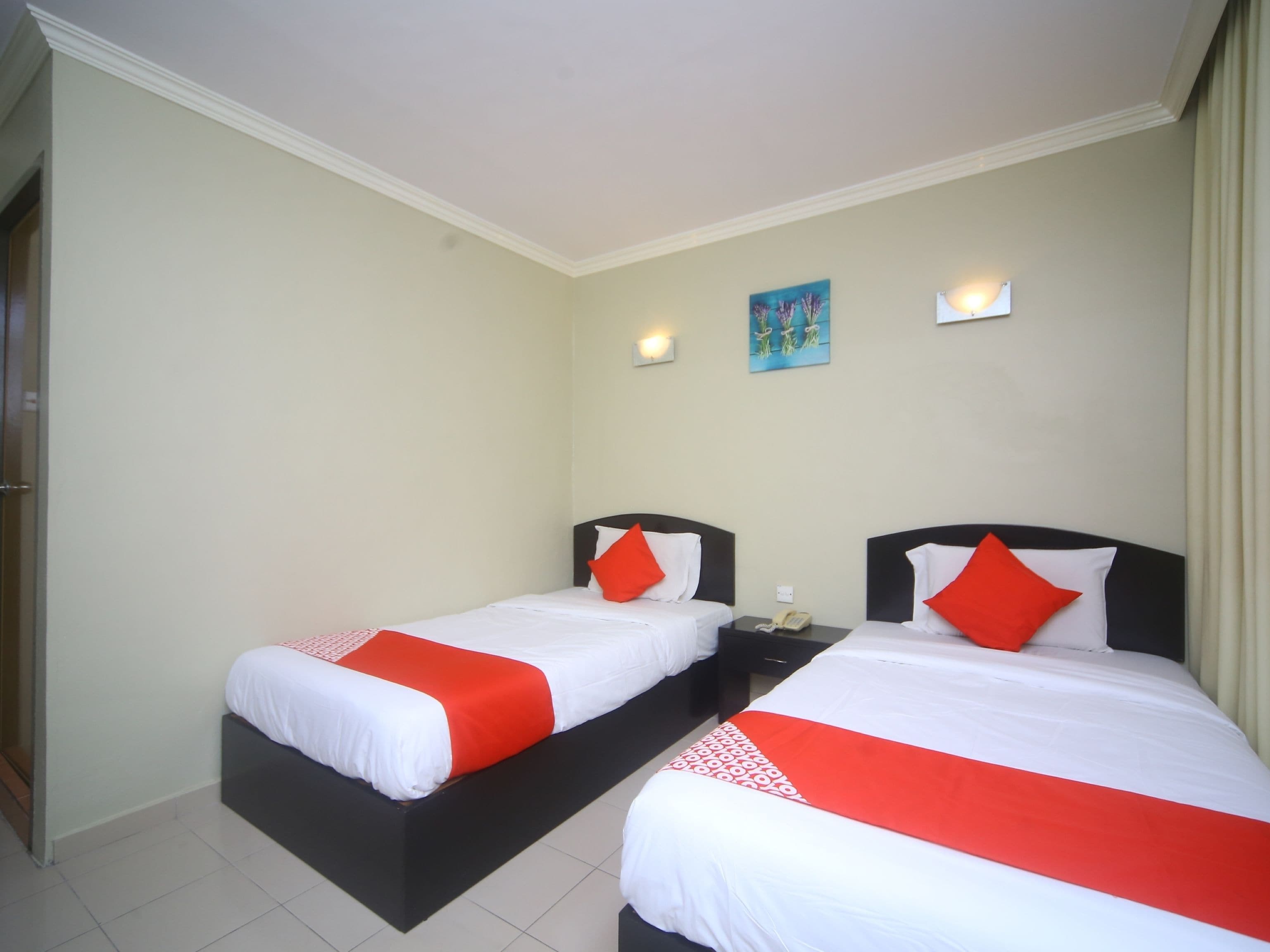 A hotel room with two beds, each with white bedding and red accents, a small bedside table with a phone, and a painting on the wall.