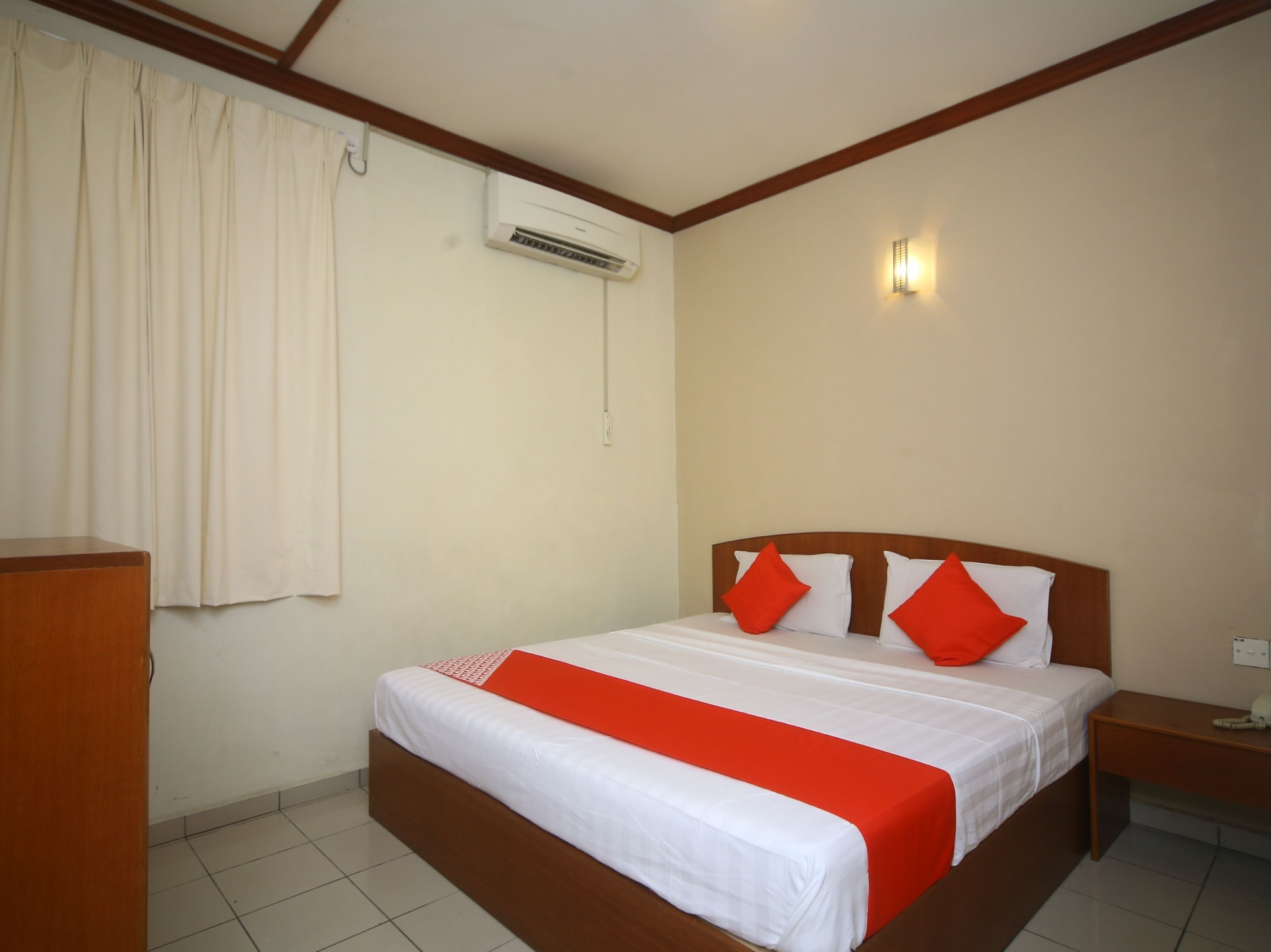 A hotel room with a bed, a nightstand, an air conditioning unit, and a wall-mounted light.