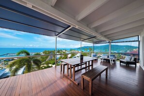 A spacious balcony with a wooden floor, a table, and seating area with a view of the sea and distant landscape. - Nautilus On The Hill - Luxury Holiday Home in Central Airlie (Airlie Beach)