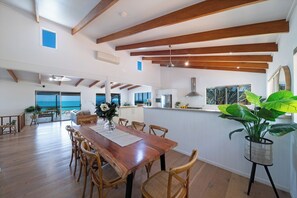 A spacious dining area with a wooden table and chairs, a kitchen with a view, and a large potted plant. - Nautilus On The Hill - Luxury Holiday Home in Central Airlie (Airlie Beach)