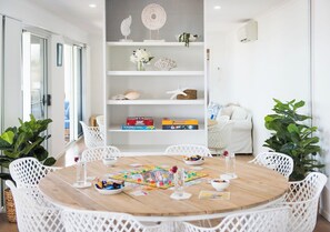 A round wooden table with white chairs, surrounded by shelves with decorative items and board games. - Nautilus On The Hill - Luxury Holiday Home in Central Airlie (Airlie Beach)