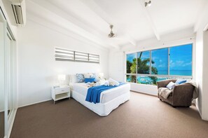 A bedroom with a large window, a bed, a nightstand, a chair, and a ceiling fan. - Nautilus On The Hill - Luxury Holiday Home in Central Airlie (Airlie Beach)