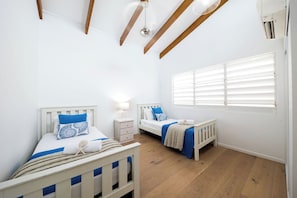 A bedroom with two beds, a window with blinds, a ceiling fan, and a wall-mounted air conditioning unit. - Nautilus On The Hill - Luxury Holiday Home in Central Airlie (Airlie Beach)