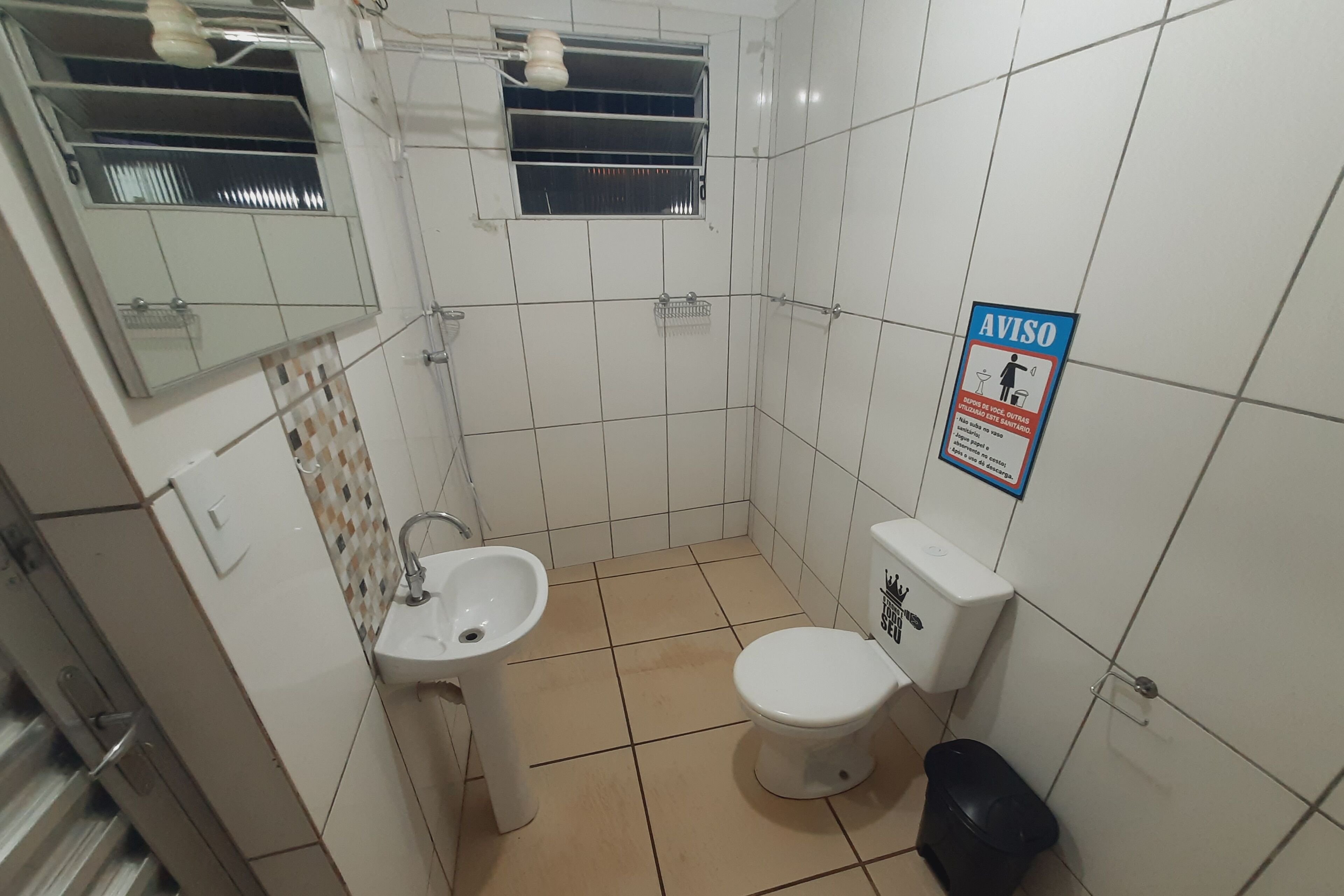 Een kleine badkamer met een wastafel, toilet en raam. Er hangt een bord aan de muur en er staat een afvalbak.