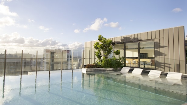 Ein Infinity-Pool auf dem Dach mit klarem Wasser, umgeben von moderner Architektur und einer Liegebereich mit weißen Liegestühlen.