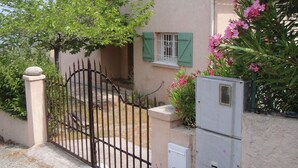 Una casa con un portón de metal, una ventana pequeña con contraventanas verdes y un arbusto de flores.