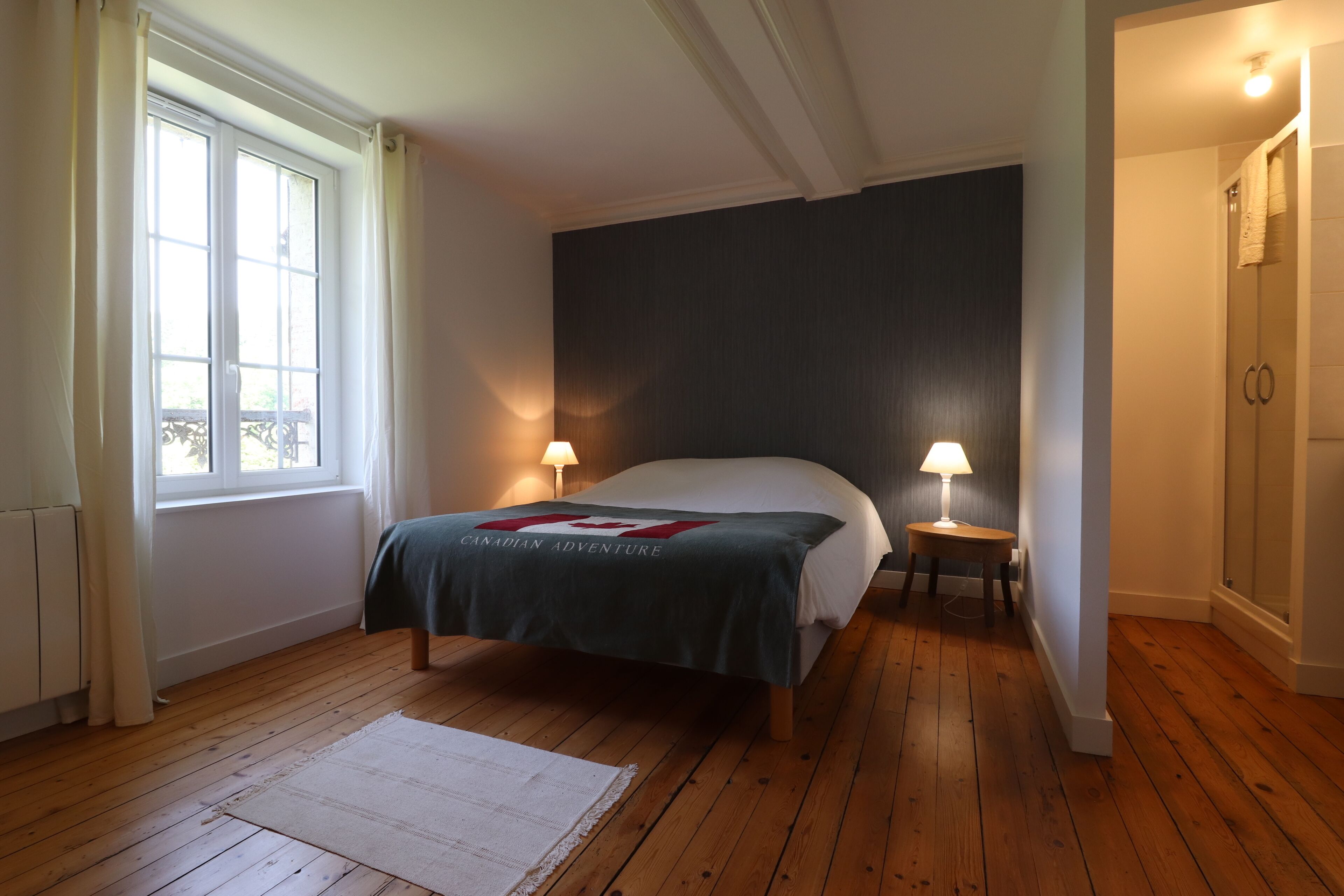 A bedroom with a wooden floor, a bed with a dark headboard, two bedside lamps, a window with curtains, and a radiator.
