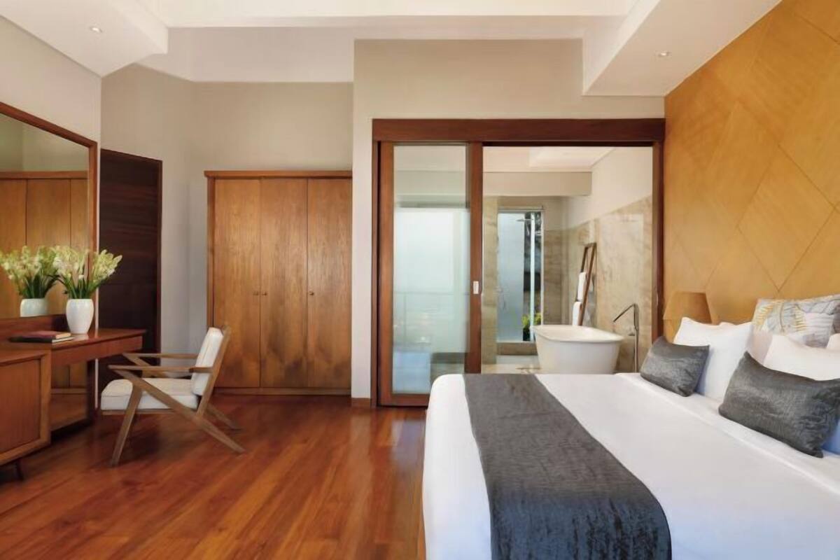 A modern hotel room with a large bed, wooden furniture, a desk, and a bathroom visible through a glass door.