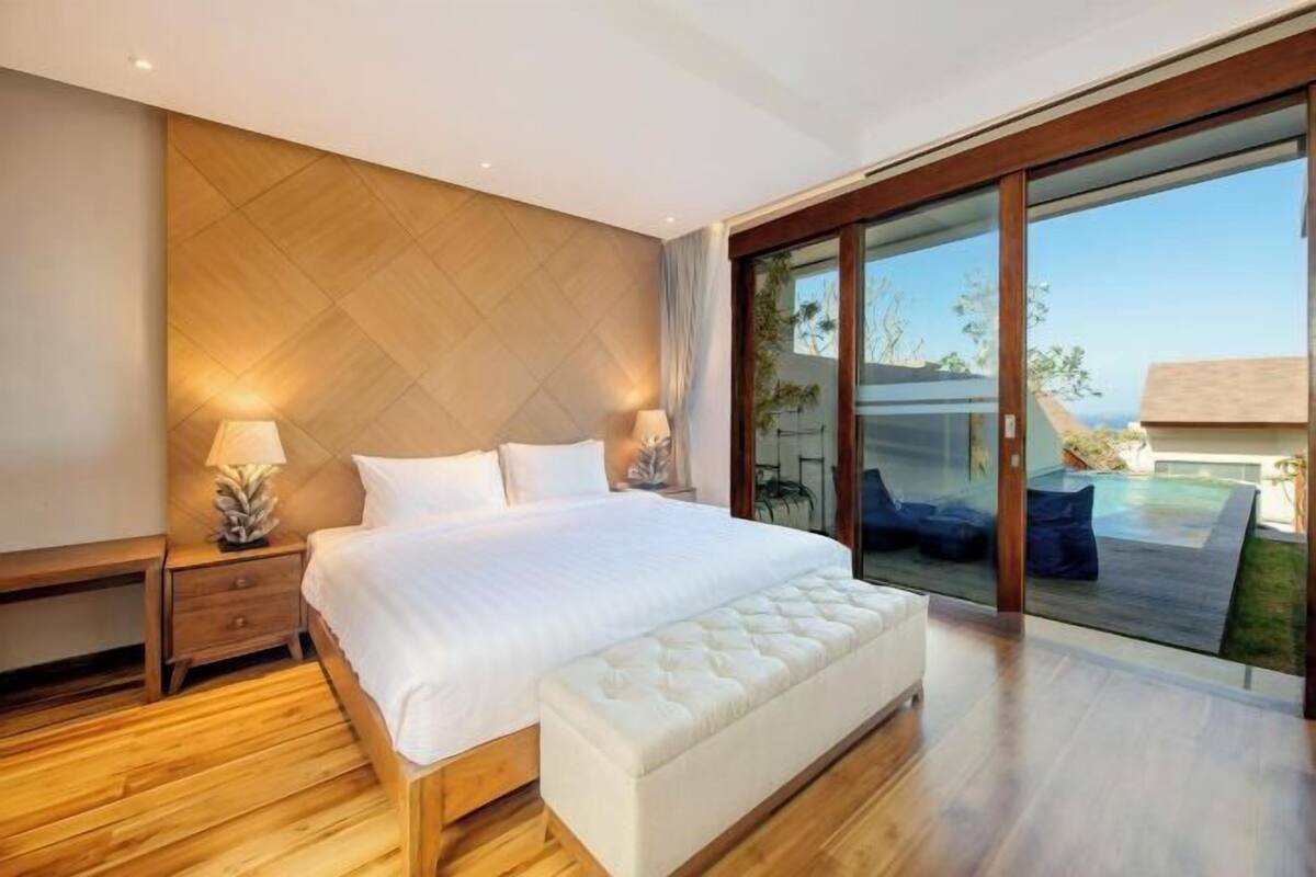 A modern bedroom with a large bed, wooden flooring, a bench, and a glass door leading to an outdoor area.