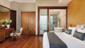Quarto de hotel moderno com piso de madeira, uma cama grande com roupa de cama cinza, uma escrivaninha com cadeira, um guarda-roupa de madeira e um banheiro com banheira e espelho.
