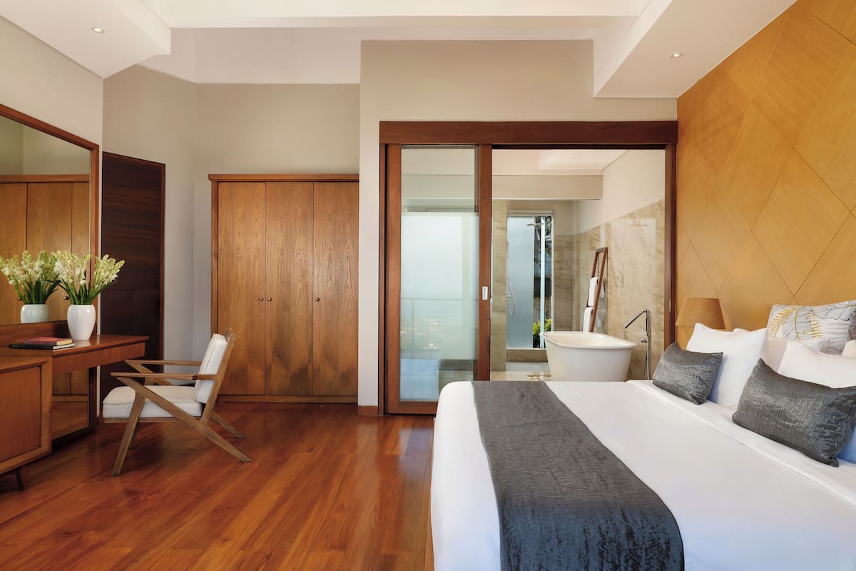 A modern hotel room with a wooden floor, a large bed with grey bedding, a desk with a chair, a wooden wardrobe, and a bathroom with a bathtub and a mirror.