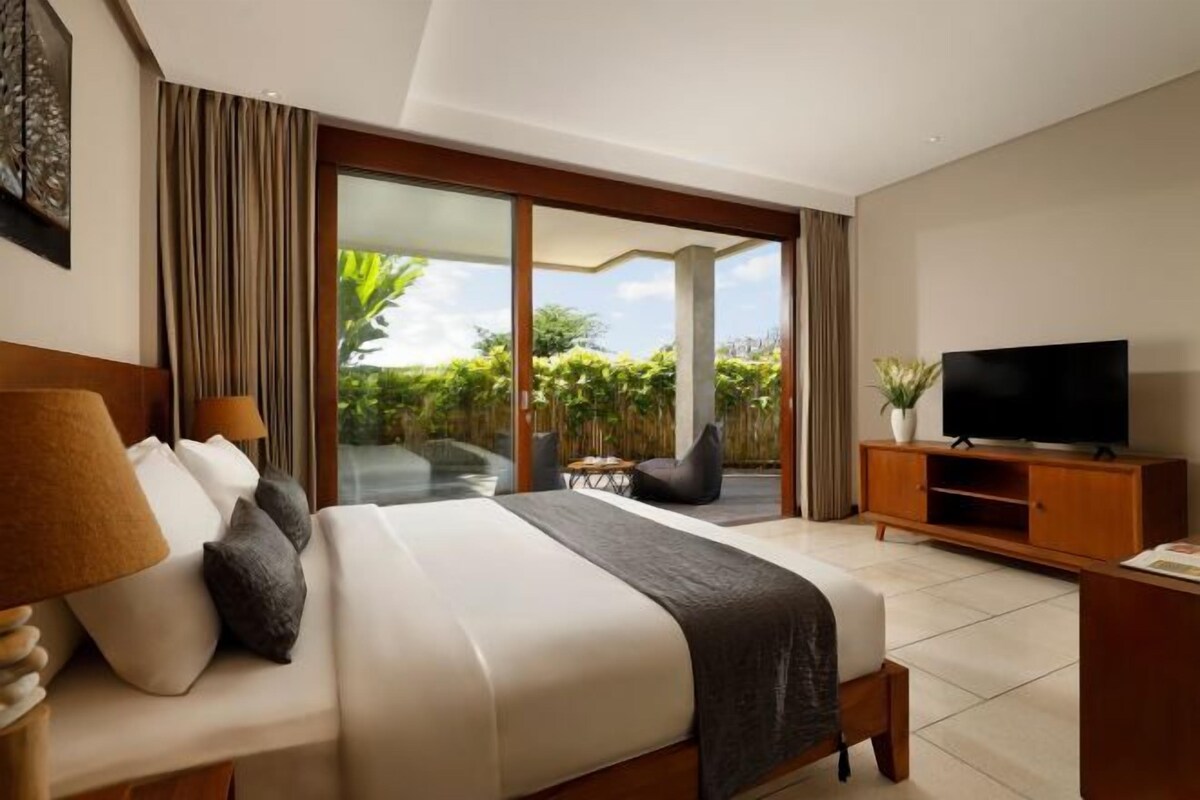 A hotel room with a large bed, a television, a sliding glass door leading to a balcony with greenery, and a small table with chairs.