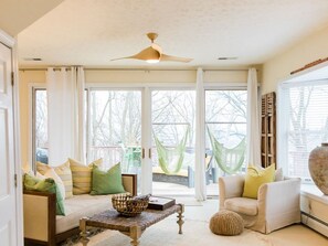 Un salon lumineux, avec un canapé, des fauteuils, une table basse et une grande fenêtre agrémentée de rideaux.
