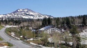 A mountain with snow-capped peaks, a road curving around a building with a sloped roof, and a forested area. - K's House Hokkaido - Asahidake Onsen Hostel (Higashikawa)