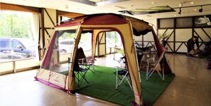 A tent set up inside a room with a green carpet, folding chairs, and a window with a view of a parked car outside. - K's House Hokkaido - Asahidake Onsen Hostel (Higashikawa)