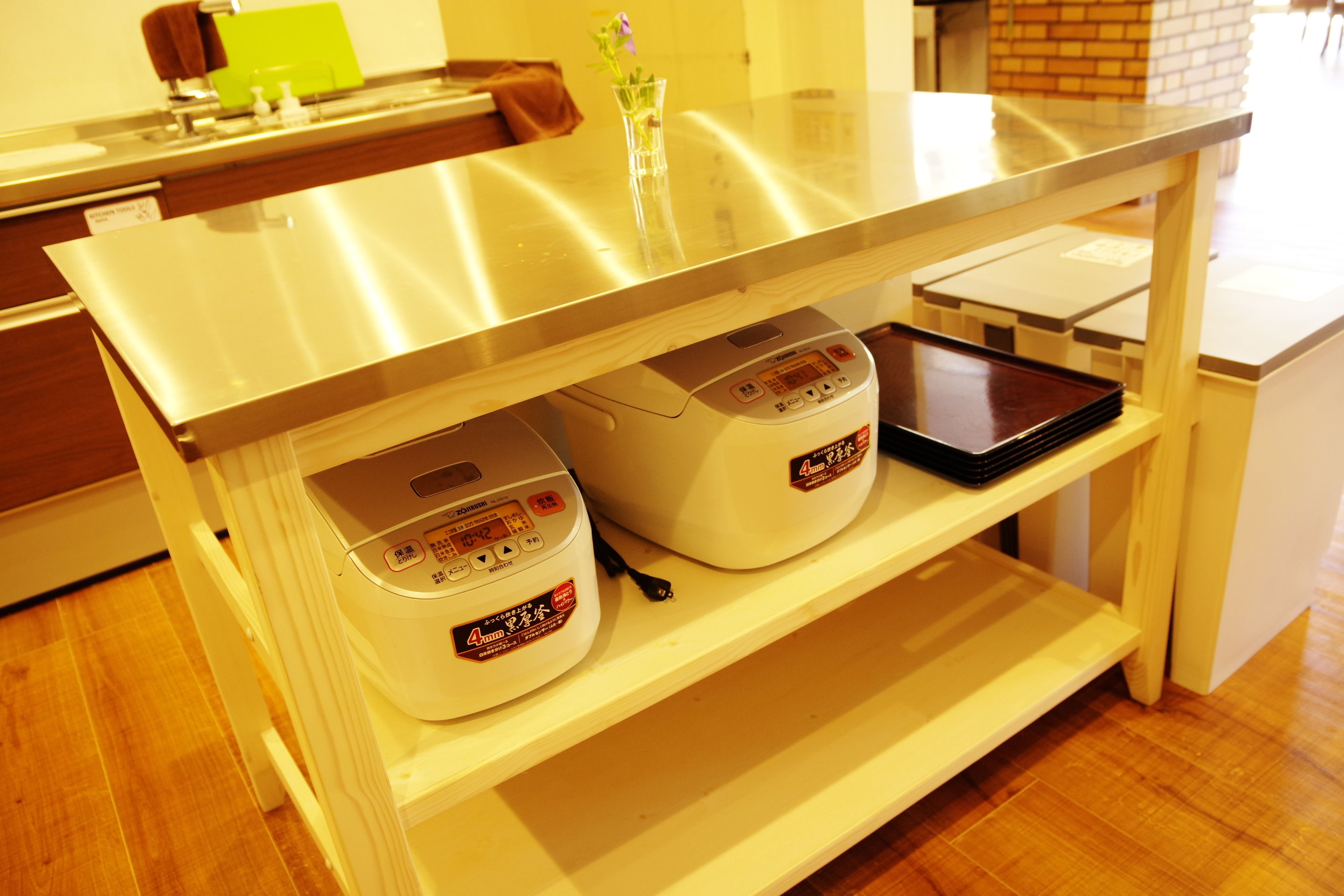 A kitchen counter with a stainless steel sink, two rice cookers, and a small vase with flowers.